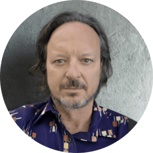 A middle aged white man faces the camera, his brown hair is pushed away from his face, he has a salt and pepper moustache and beard and wears a retro blue shirt.